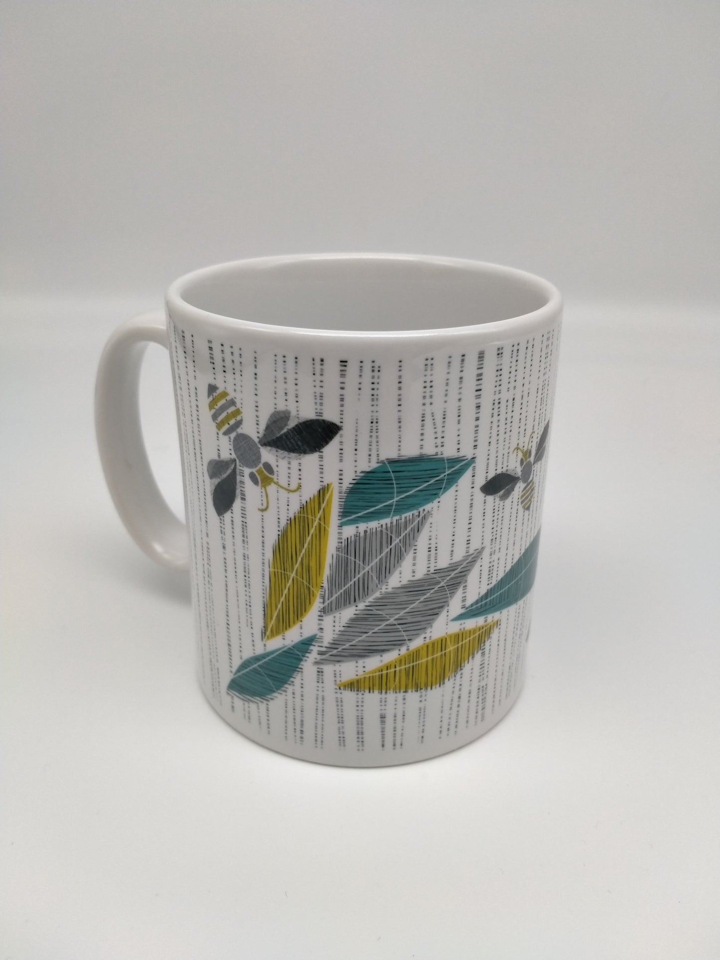White mug with leaf and bee design. Grey, black, gold and teal tones.