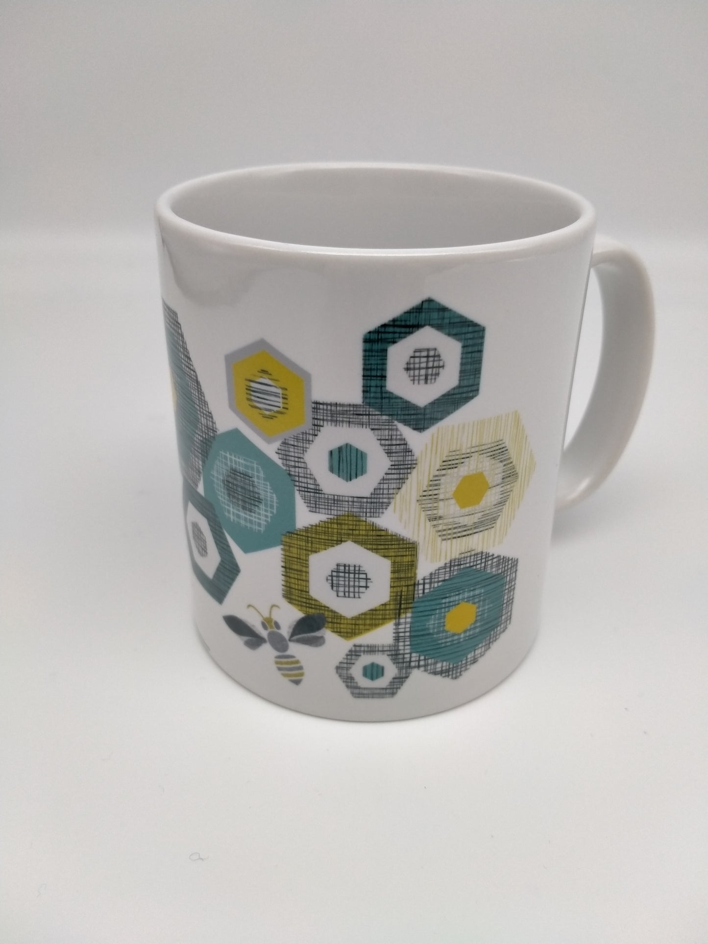 White mug with multicoloured hexagons and bee design. Grey, black, gold and teal tones.