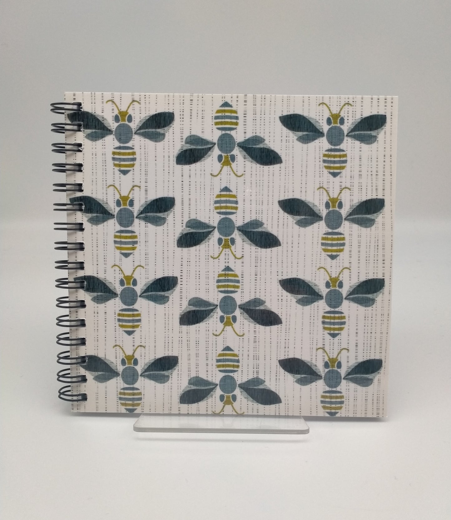 A square notebook with 12 bees in three rows on the cover. Spiral bound. Grey, black and gold tones.