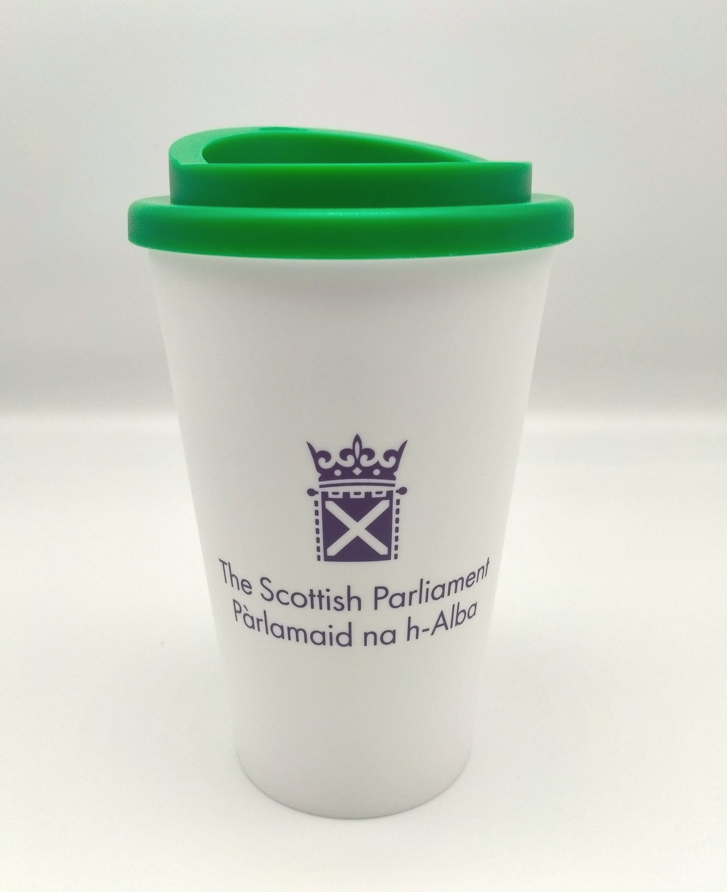 A white cup with a green lid, decorated with the symbol of the Scottish Parliament in purple.