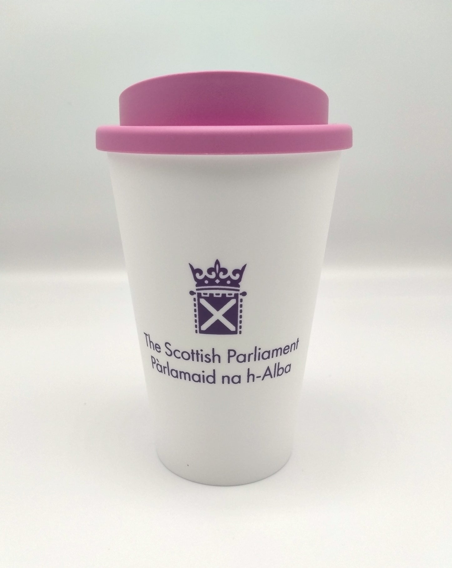 A white cup with a pink lid, decorated with the symbol of the Scottish Parliament in purple.