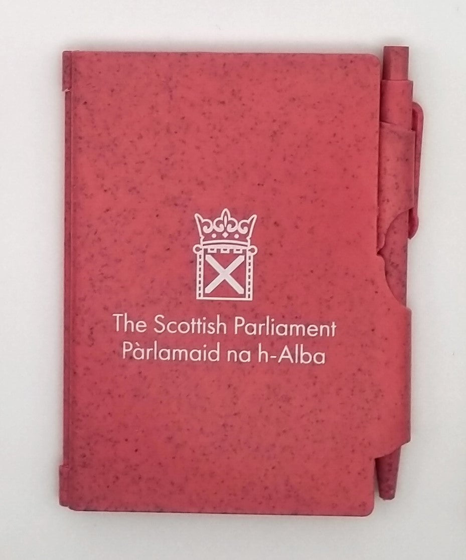 A reddish-pink notebook with the symbol of the Scottish Parlaiment in white and a matching pen slid into a holder on the side.