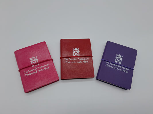 Three small notebooks in pink, red and purple decorated with the symbol of the Scottish Parliament.