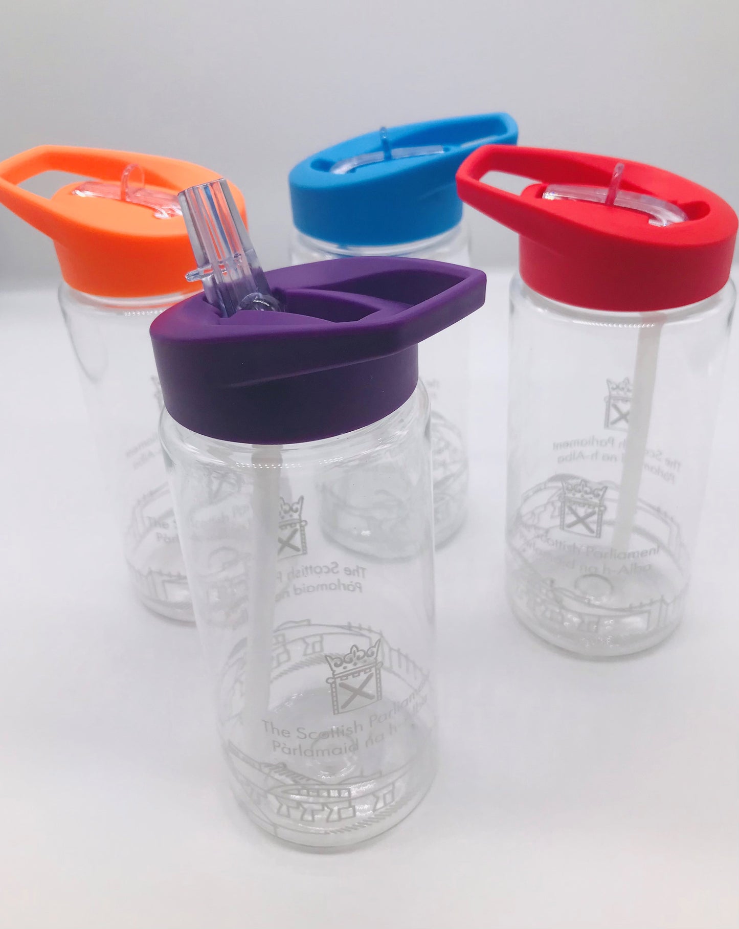 Four clear plastic bottles with internal straws. They have the Scottish Parliament logo and outline of the parliament building painted around the circumference of the bottle. Each bottle has a different coloured lid. There is orange, blue, red, and purple. Each lid has a foldable spout for drinking.