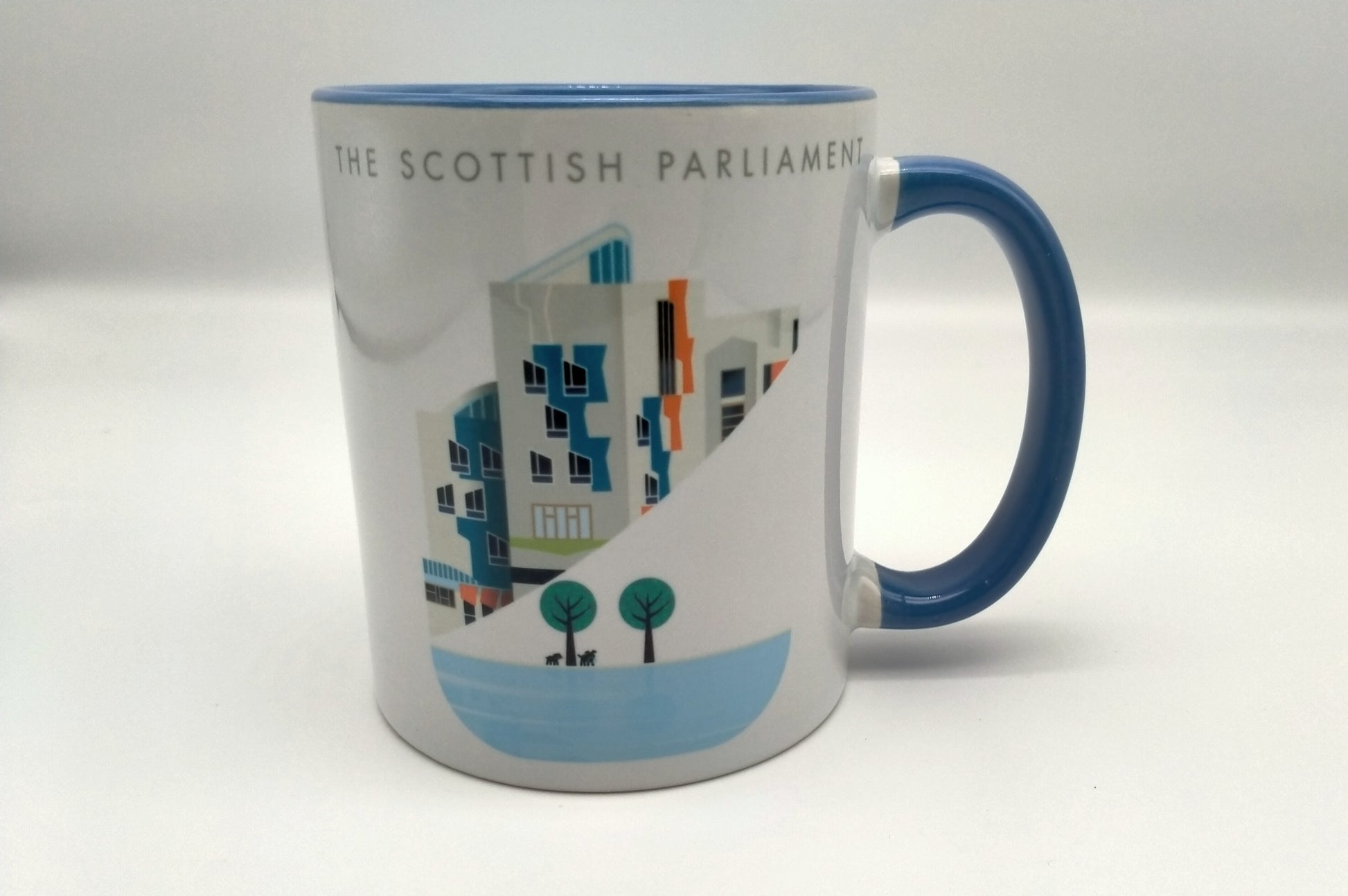 A white mug printed with an image of a part of the Scottish Parliament building. A contemporary design features some architectural details of the building, a pond and trees. Writing The Scottish Parliament in English. Blue handle.