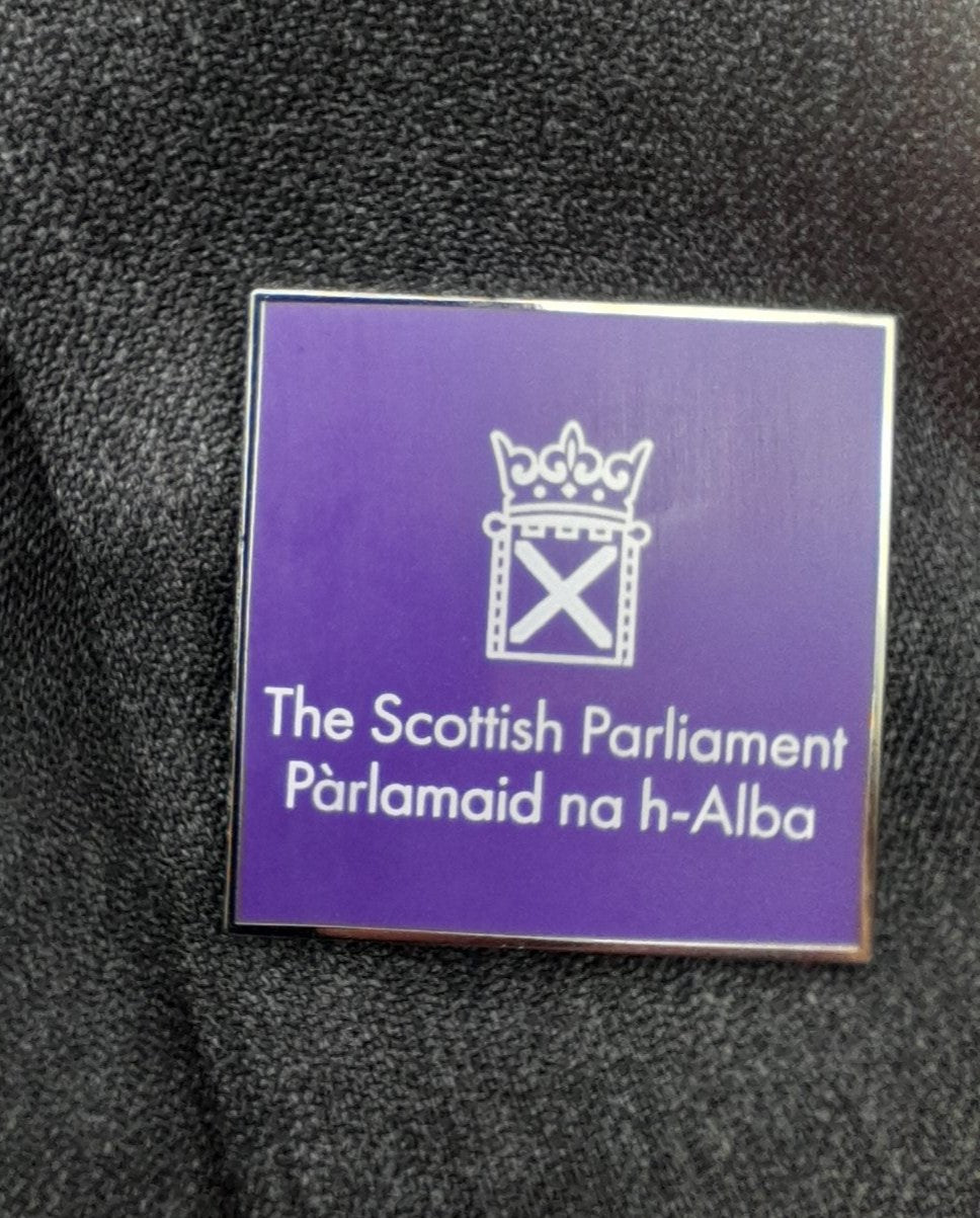 Square purple badge with the white symbol of the Scottish Parliament pinned to grey fabic.