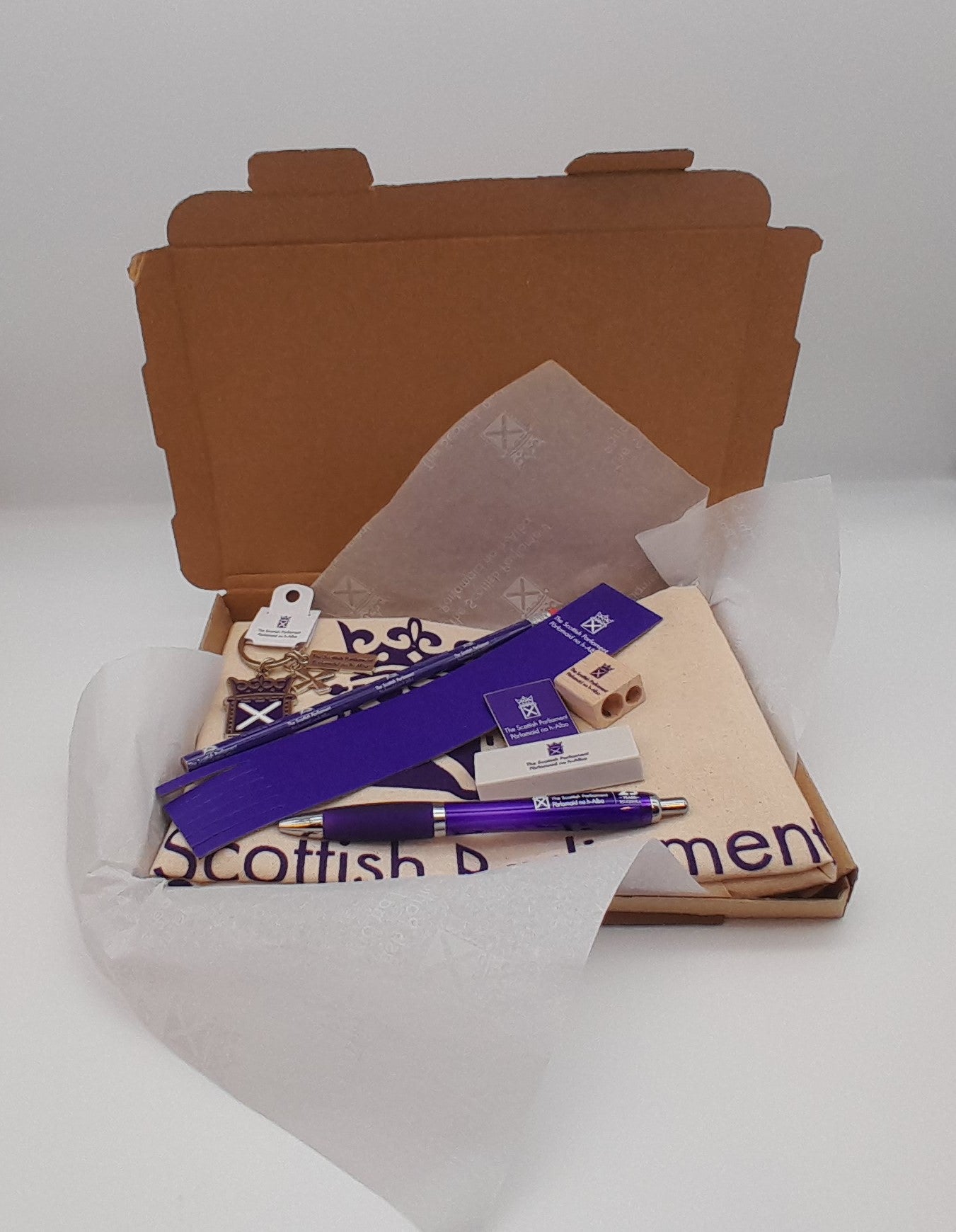 An open box lined with tissue paper, containing a cotton shopper, a pen, an eraser, a pencil, a sharpener, a bookmark, a pin badge and a keyring. 