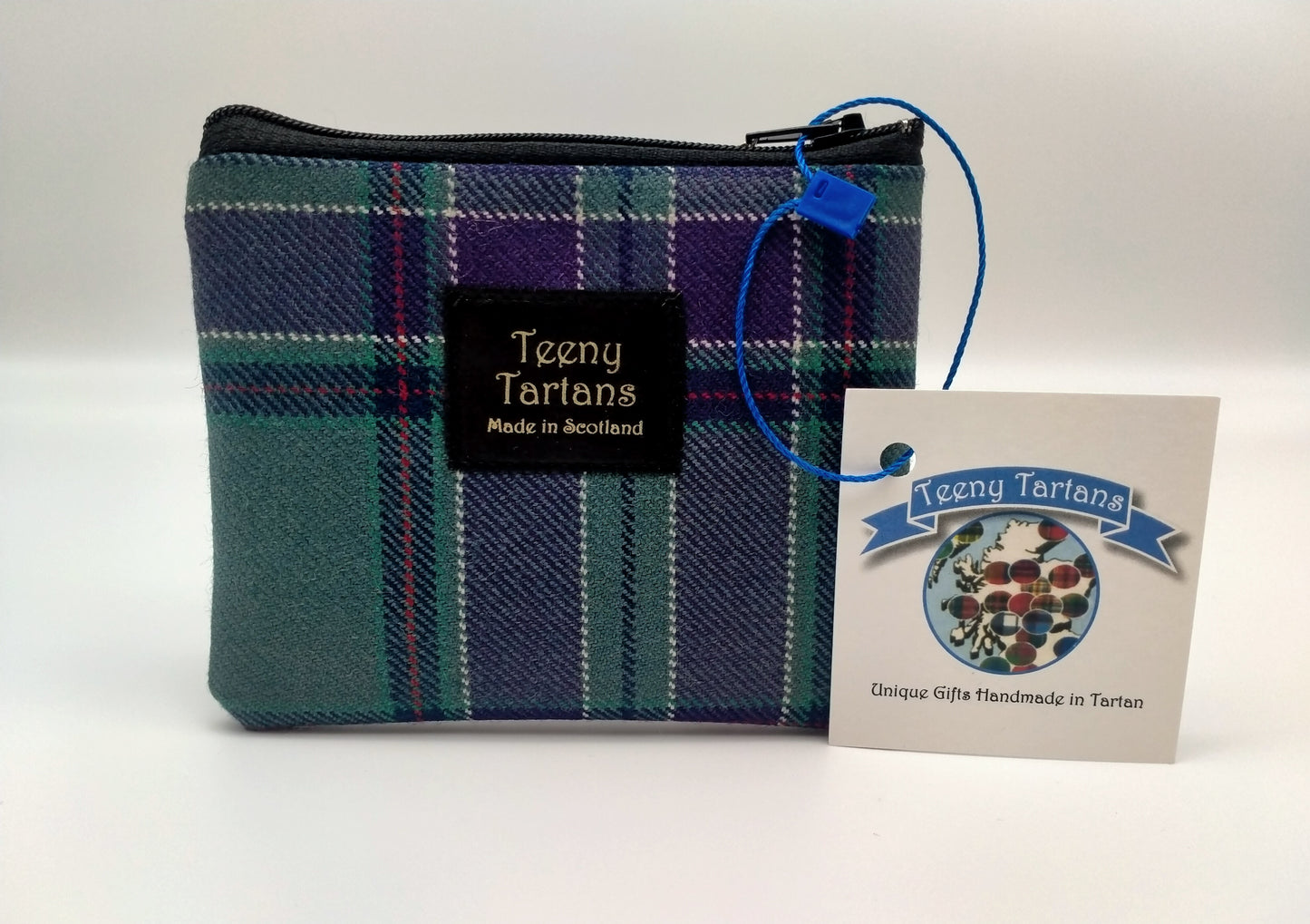 A rectangular purse made of the Scottish Parliament tartan with a black label in the middle. The writing on the lablel says: Teeny Tartans. Made in Scotland. A brand tag attached to the side.