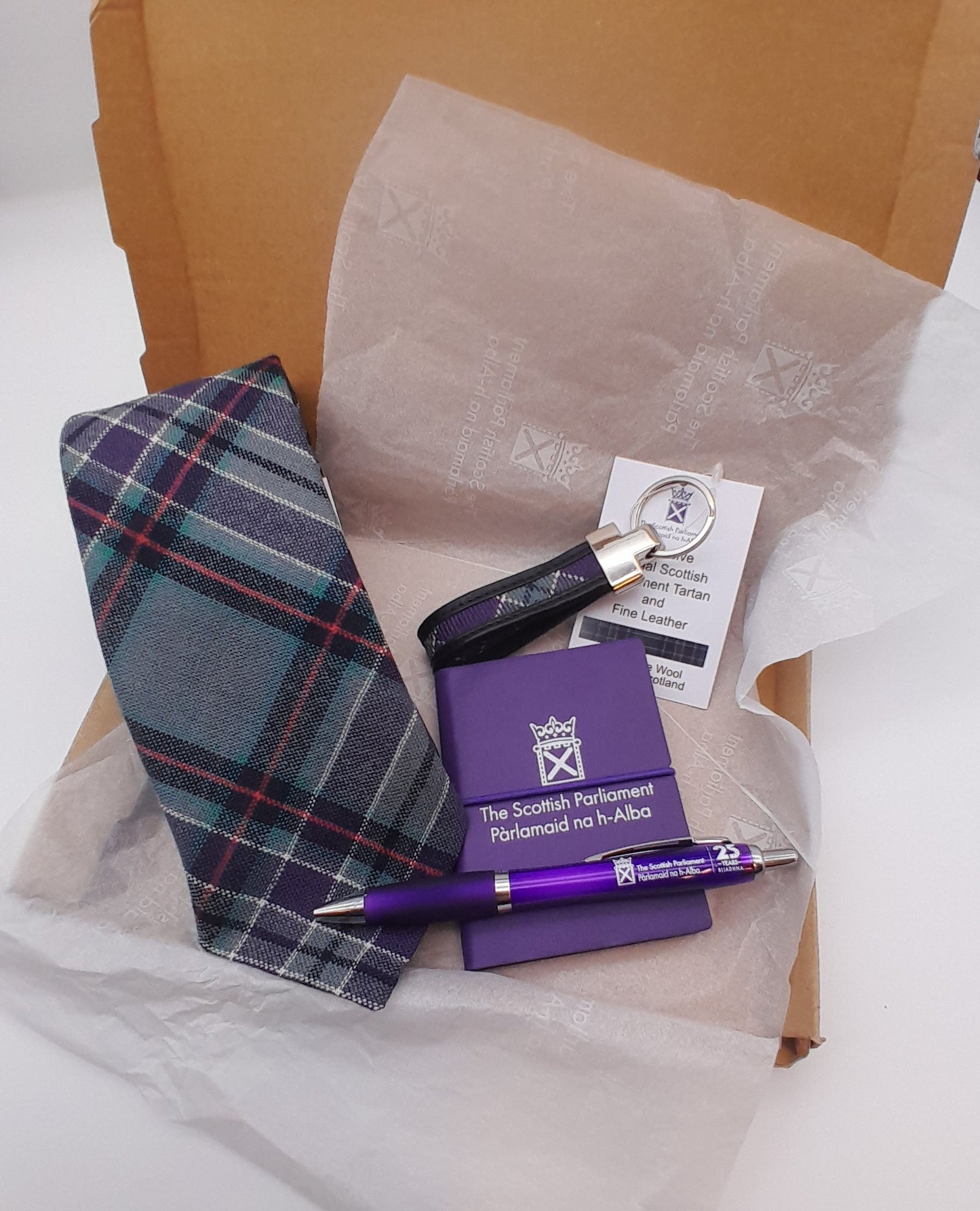 A open box with white tissue lining, a folded tartan tie, a keyring, a notebook and a pen. 