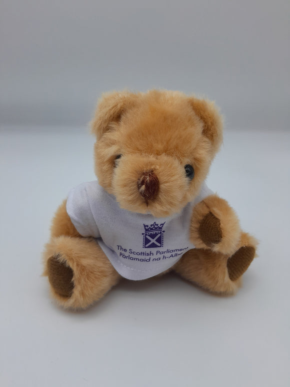 A small soft toy bear. Light brown, wearing a white t-shirt with the symbol of the Scottish Parliament in purple. 