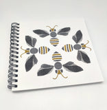A square notebook with four bees on the cover. Spiral bound. Grey, black and gold tones.
