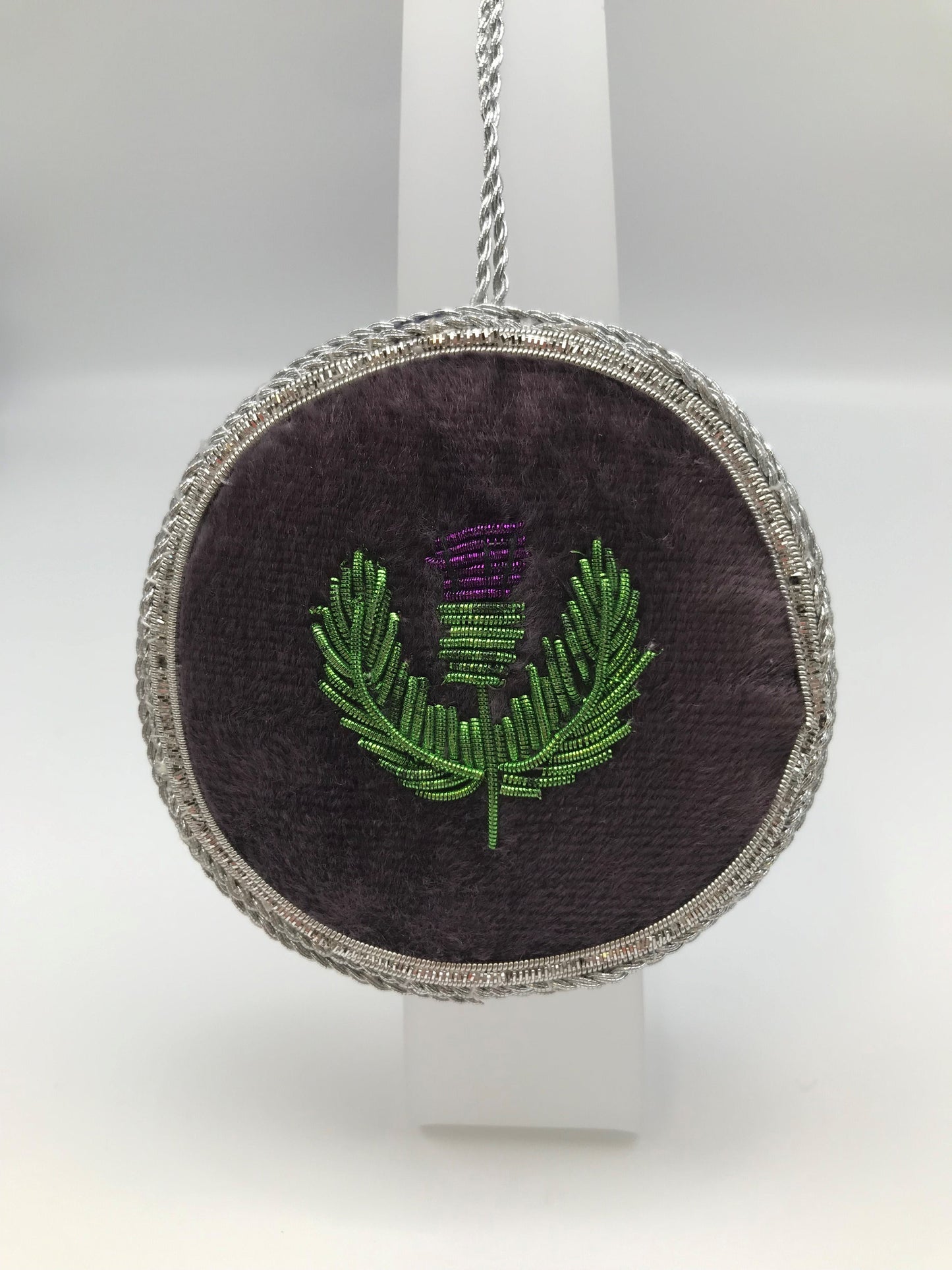 Round decoration with thistle embroidered over purple fabric and silver stitching detail.