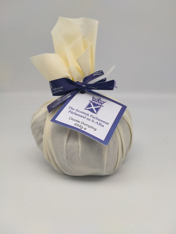 A round cake wrapped in linen tied with a purple ribbon. An attached label shows the symbol of the Scottish Parliament and reads: 'Clootie Dumpling, 450g'.