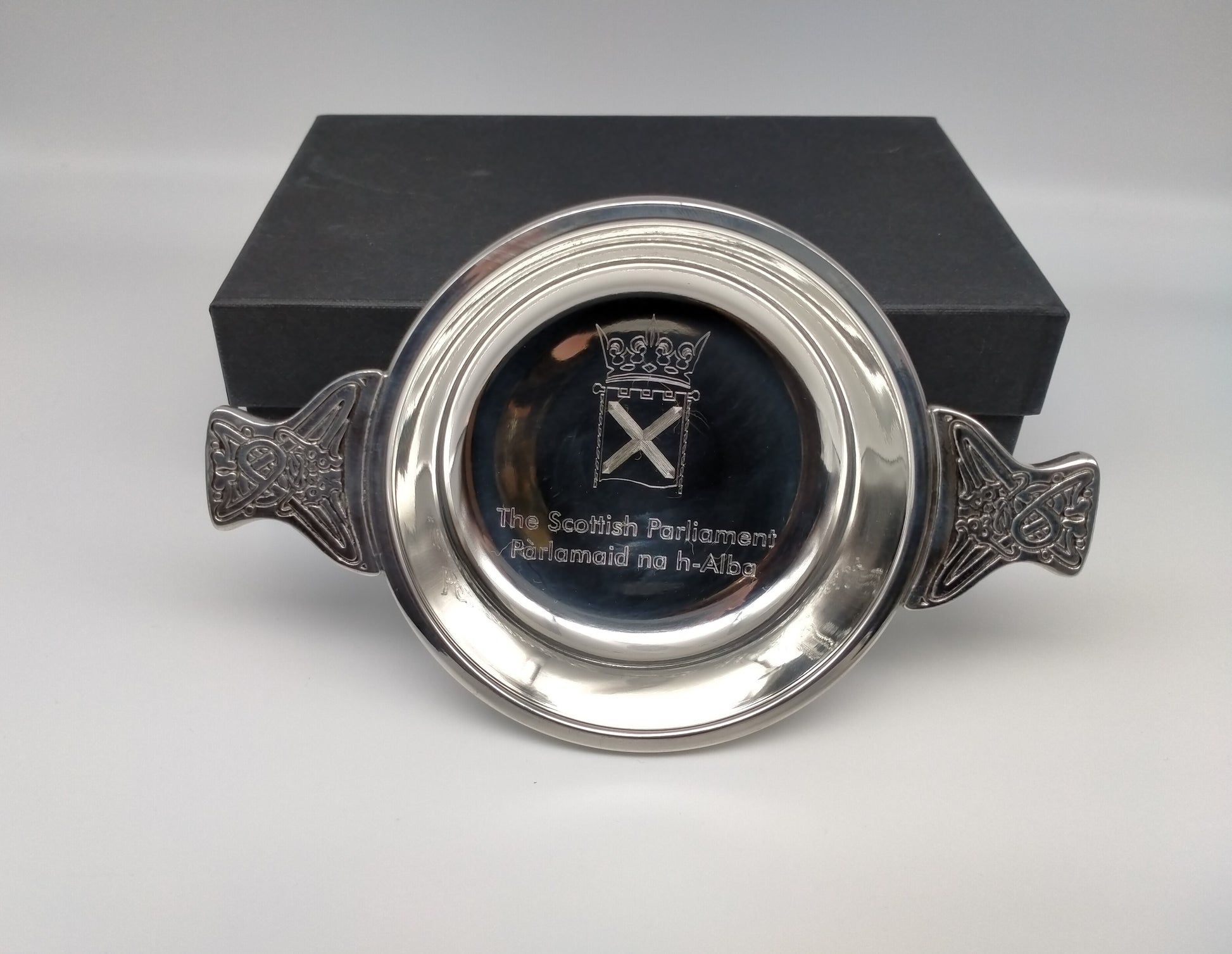 Silver coloured round quaich with handles featuring a Celtic knot design. Bowl engraved with the Parliament symbol. Presented next to black box.