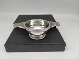 Silver coloured round quaich with handles featuring a Celtic knot design. Bowl engraved with the Parliament symbol. Presented next to black box.