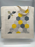 A square cotton shopping bag printed with an image of a bee and honeycomb in grey, black and gold on off-white background.