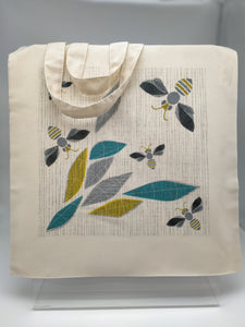 A square cotton shopping bag printed with an image of a bee and honeycomb in grey, black and gold on off-white background.