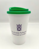 A white cup with a green lid, decorated with the symbol of the Scottish Parliament in purple.