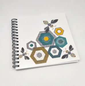 A square notebook with bees and leaf shapes on the cover. Spiral bound. Grey, black and gold tones.