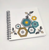 A square notebook with bees and hexagon shapes on the cover. Spiral bound. Grey, black and gold tones.