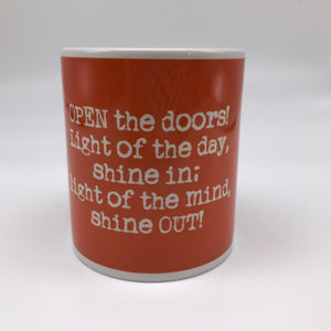 An orange mug with writing in white. The words read: Open the doors! Light of the day, shine in; light of the mind, shine out!.