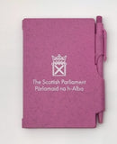 A pink notebook with the symbol of the Scottish Parlaiment in white and a matching pen slid into a holder on the side.