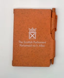 An orange notebook with the symbol of the Scottish Parlaiment in white and an orange pen slid into a holder on the side.